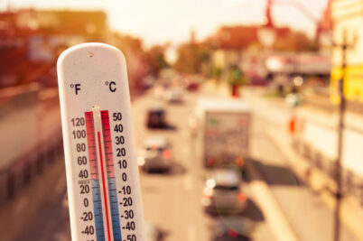 A thermometer showing 102 degrees Fahrenheit with urban traffic and buildings in the background